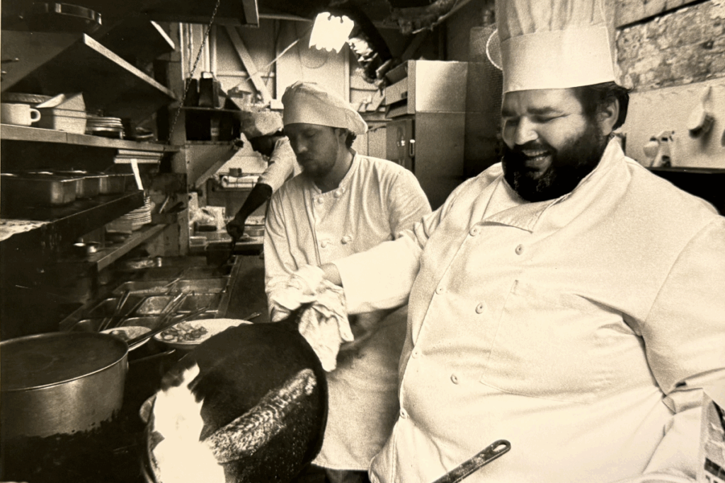 Two chefs in a bustling kitchen, one smiling broadly and the other focused on cooking. The atmosphere is warm and lively, with stainless steel cookware.