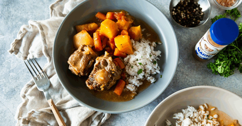 A hearty bowl of oxtail stew with tender meat, potatoes, carrots, and rice. Surrounded by a fork, spices, and fresh parsley on a textured surface.