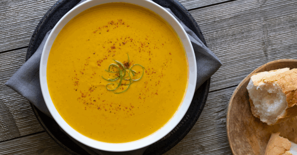 A bowl of creamy orange soup sprinkled with paprika and garnished with green curls on a wooden table. Nearby, bread sits in a small bowl.