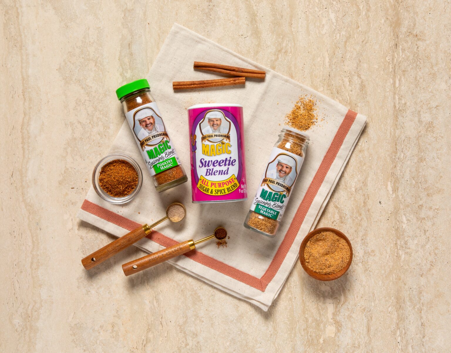 Three Chef Paul Prudhomme's Magic seasoning products arranged on a cream linen napkin with yellow and orange flowers - two glass spice jars (Vegetable Magic and Poultry Magic) and one pink container of Sweetie Blend sugar and spice blend.