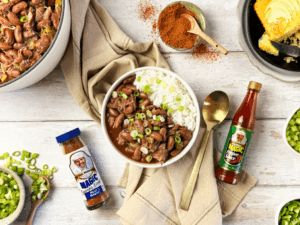 A bowl of red beans and rice topped with green onions, surrounded by cornbread, seasonings, hot sauce, and a pot of beans.