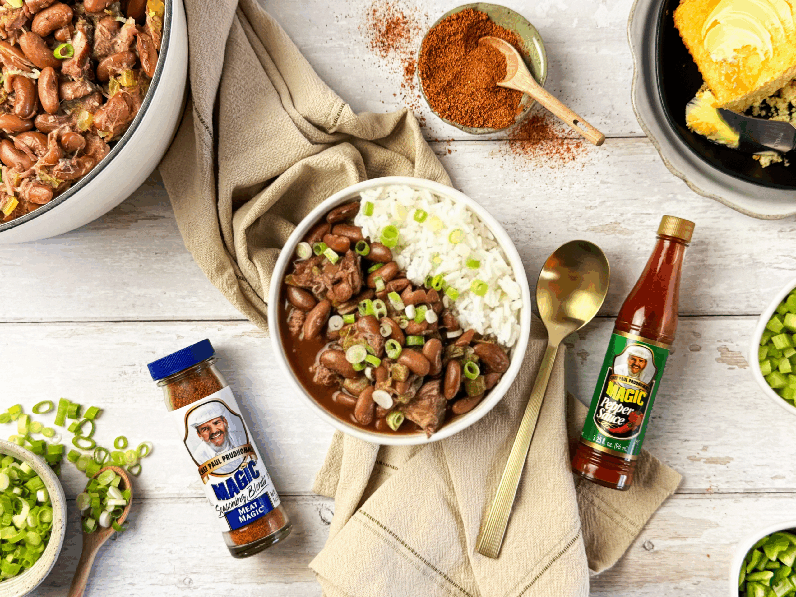 A bowl of red beans and rice topped with green onions, surrounded by cornbread, seasonings, hot sauce, and a pot of beans.