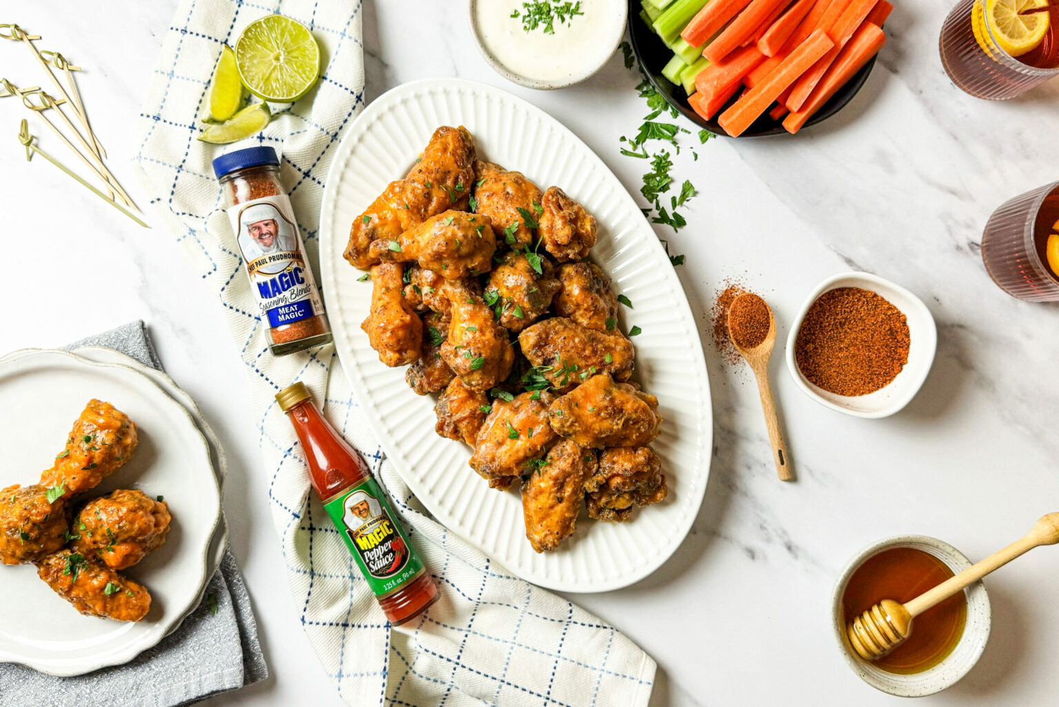 A platter of honey buffalo chicken wings garnished with herbs, served with celery, carrot sticks, dipping sauce, seasonings, hot sauce, and drinks with lemon slices.
