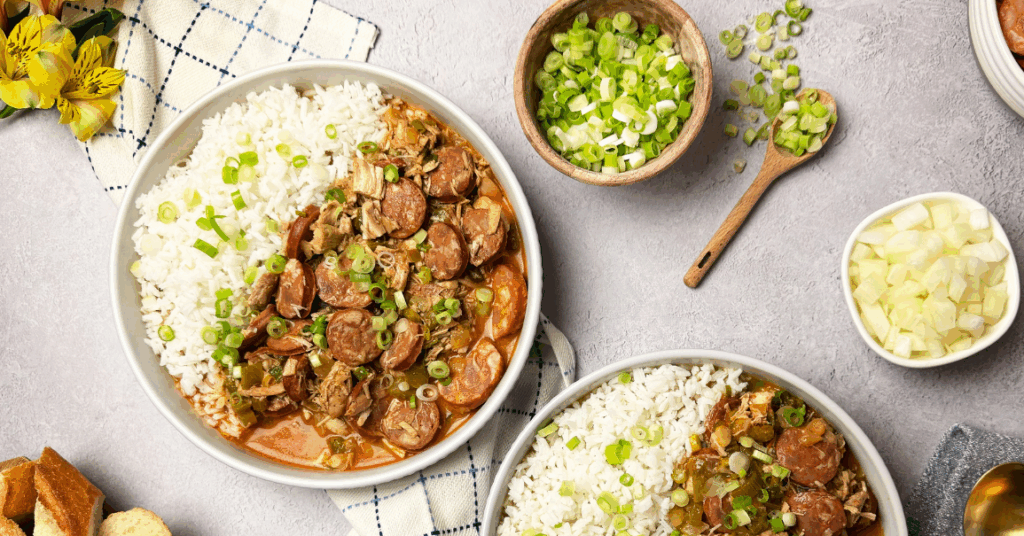 Two bowls of chicken and sausage gumbo with rice, topped with green onions, surrounded by sliced bread, sausage, chopped vegetables, and gold spoons on a gray napkin.