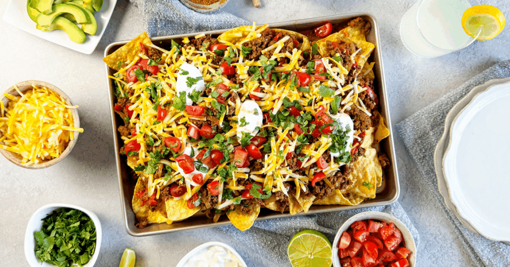 A tray of loaded nachos topped with beef, cheese, tomatoes, sour cream, and cilantro, surrounded by bowls of salsa, shredded cheese, avocado, tomatoes, lime wedges, and drinks with lemon slices.