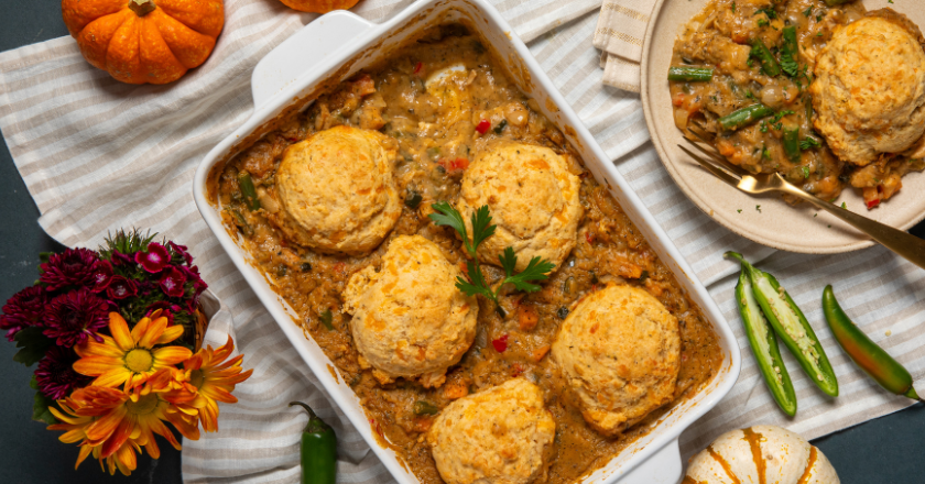 A casserole dish with golden biscuits on a stew-like base is on a striped cloth. Surrounded by a spice jar, green chilies, mini pumpkins, flowers, and plates. Cozy and autumnal.