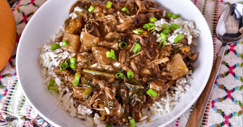 A bowl of savory beef stew with chunks of meat and vegetables sits over white rice, garnished with chopped green onions. The setting is bright and colorful.