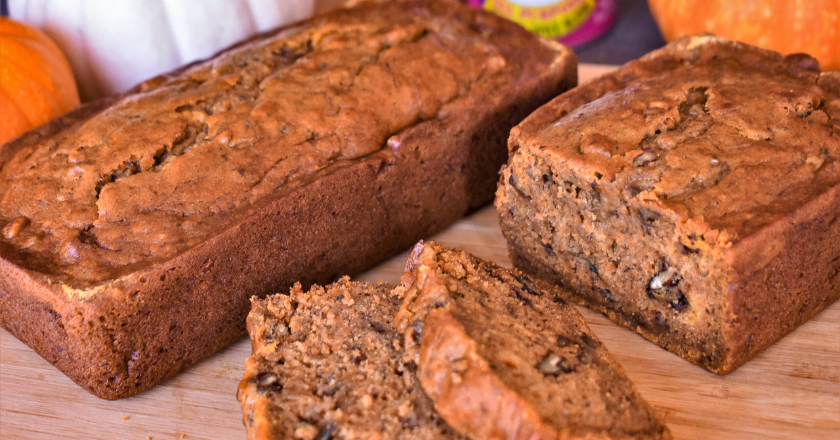 Close-up of two loaves of pumpkin bread on a wooden board, surrounded by small pumpkins. One loaf is sliced, revealing a moist, rich texture.