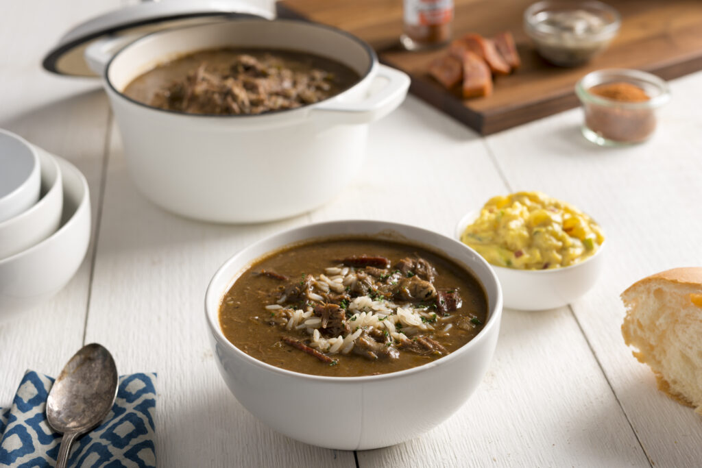 Gumbo in a white bowl on a white wooden table next to a large pot of gumbo