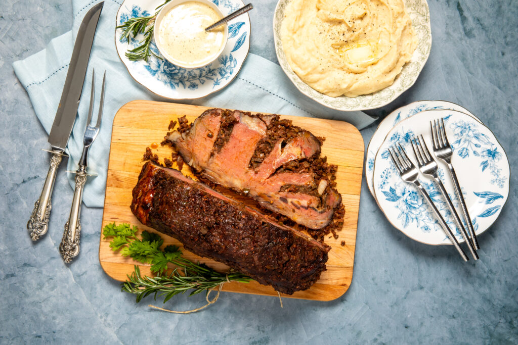 Ribeye roast on a cutting board showing a cross-section cut of meat. Next to a bowl of mashed potatoes, silver serving utensils and decorative plates on a blue background.