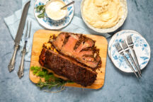 Ribeye roast on a cutting board showing a cross-section cut of meat. Next to a bowl of mashed potatoes, silver serving utensils and decorative plates on a blue background.