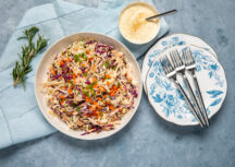 a white serving bowl filled with shredded green cabbage, purple cabbage, and carrots. sprinkled with green onions. on a blue background with a light blue napkin and serving plates with forks