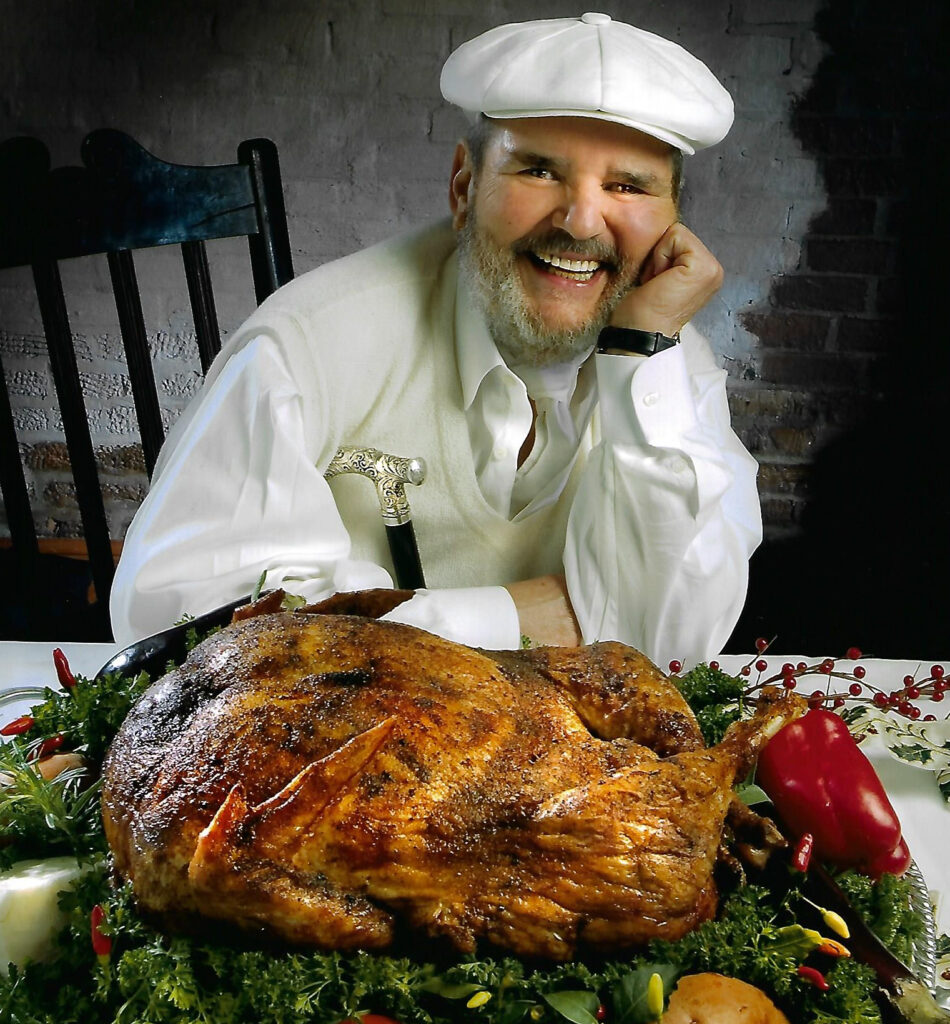 Chef Paul smiling in a white cap and shirt sits at a table with a golden roasted turkey garnished with herbs and peppers, conveying warmth and celebration.