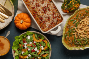 Thanksgiving spread with a vibrant salad topped with pomegranate and goat cheese, a casserole with breadcrumbs, and a green bean dish, surrounded by small pumpkins.