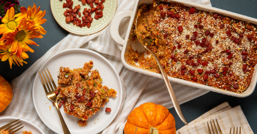 A baked dish garnished with bacon sits beside a plate with a serving, surrounded by orange flowers and small pumpkins. Warm, autumnal ambiance.