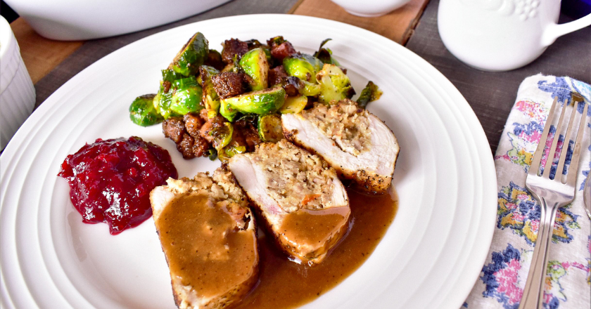 A white plate with sliced stuffed turkey topped with gravy, roasted Brussels sprouts and bacon, and a serving of cranberry sauce. A floral napkin and fork sit beside the plate.