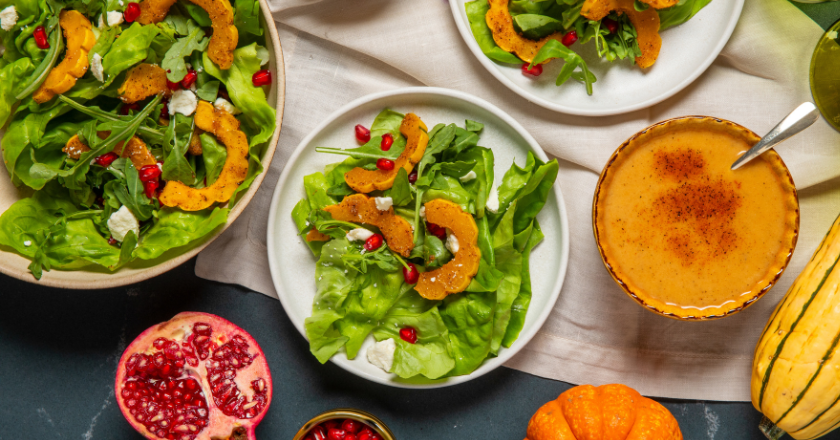 A vibrant fall salad spread features mixed greens, roasted squash, pomegranate seeds, and feta cheese. Accompanied by creamy soup, a halved pomegranate, and small pumpkins, it conveys a warm, seasonal feel.