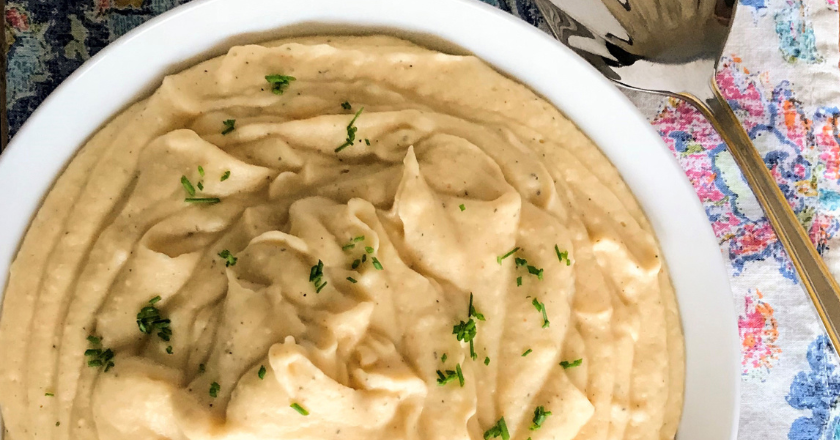 A bowl of creamy mashed potatoes garnished with chopped parsley sits on a patterned tablecloth. A shiny serving spoon is placed beside the bowl.