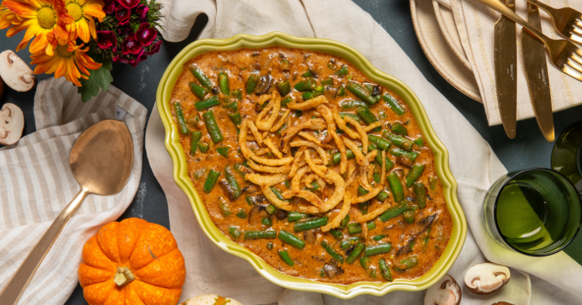 A green dish filled with green bean casserole topped with crispy fried onions, surrounded by autumnal decor, flowers, a pumpkin, and tableware. Cozy and festive.