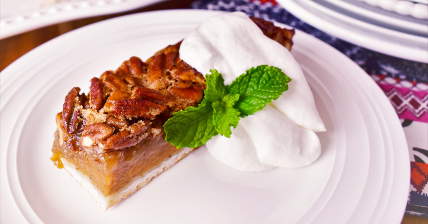 A slice of pecan pie on a white plate with fluffy whipped cream and a fresh mint garnish, set on a patterned tablecloth, creating a cozy, inviting feel.