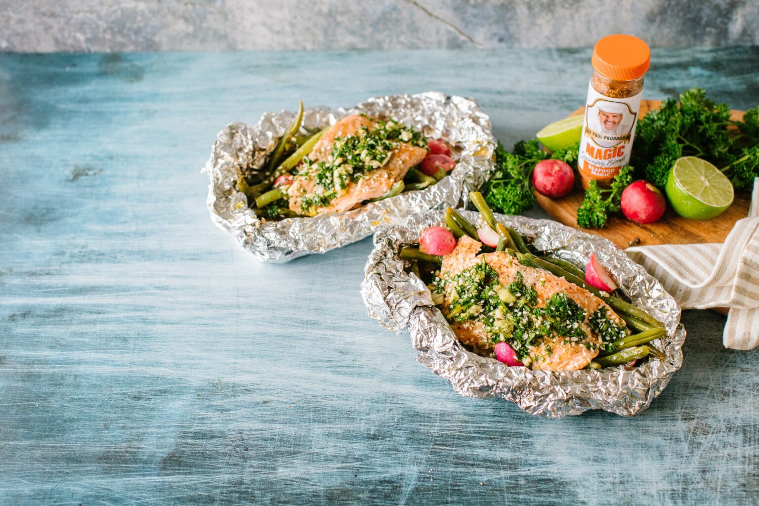 Grilled salmon with herbs, green beans, and radishes in foil on a blue table. In the background, lime, Magic Seasoning spices, and parsley add freshness.