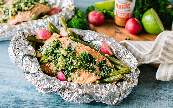 Grilled salmon with herbs, green beans, and radishes in foil on a blue table. In the background, lime, spices, and parsley add freshness.