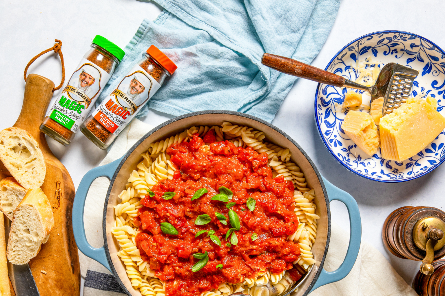 Overhead shot of rotini pasta topped with rich red tomato sauce and fresh basil, surrounded by Chef Paul Prudhomme’s Magic Poultry Magic and Seafood Magic seasoning bottles, grated Parmesan, and sliced bread.