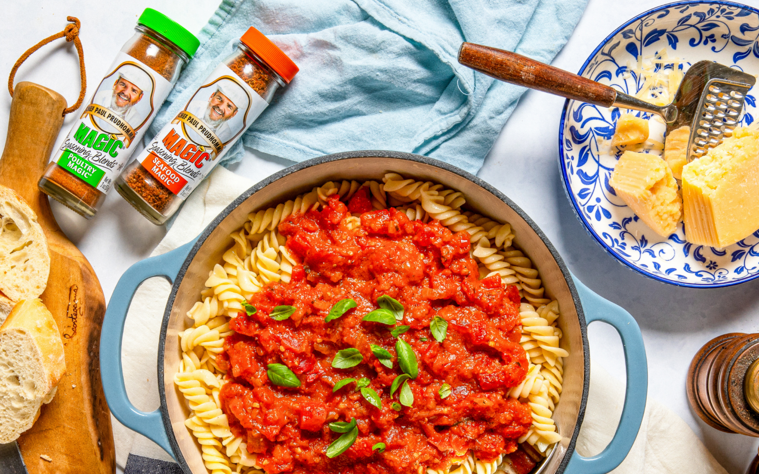 Overhead shot of rotini pasta topped with rich red tomato sauce and fresh basil, surrounded by Chef Paul Prudhomme’s Magic Poultry Magic and Seafood Magic seasoning bottles, grated Parmesan, and sliced bread.