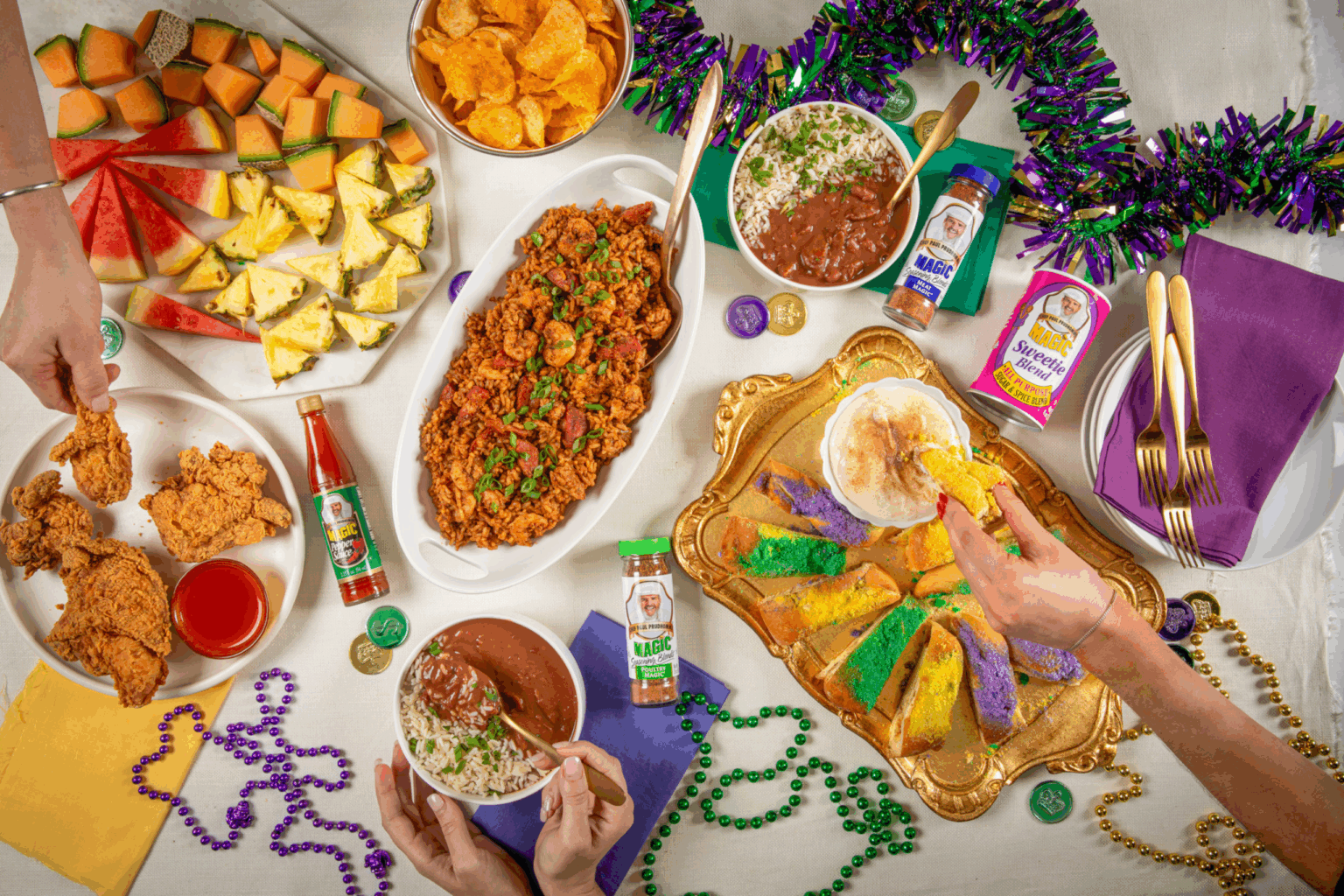 Festive table with colorful Mardi Gras decor, fried chicken, jambalaya, king cake, fruit, potato chips, hot sauce, and accessories, creating a joyful celebration.