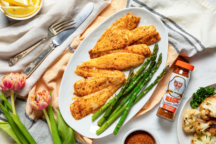 Broiled fish fillets with asparagus on a white plate, accompanied by a spice bottle. Lemons, cutlery, flowers, and cauliflower surround the scene.