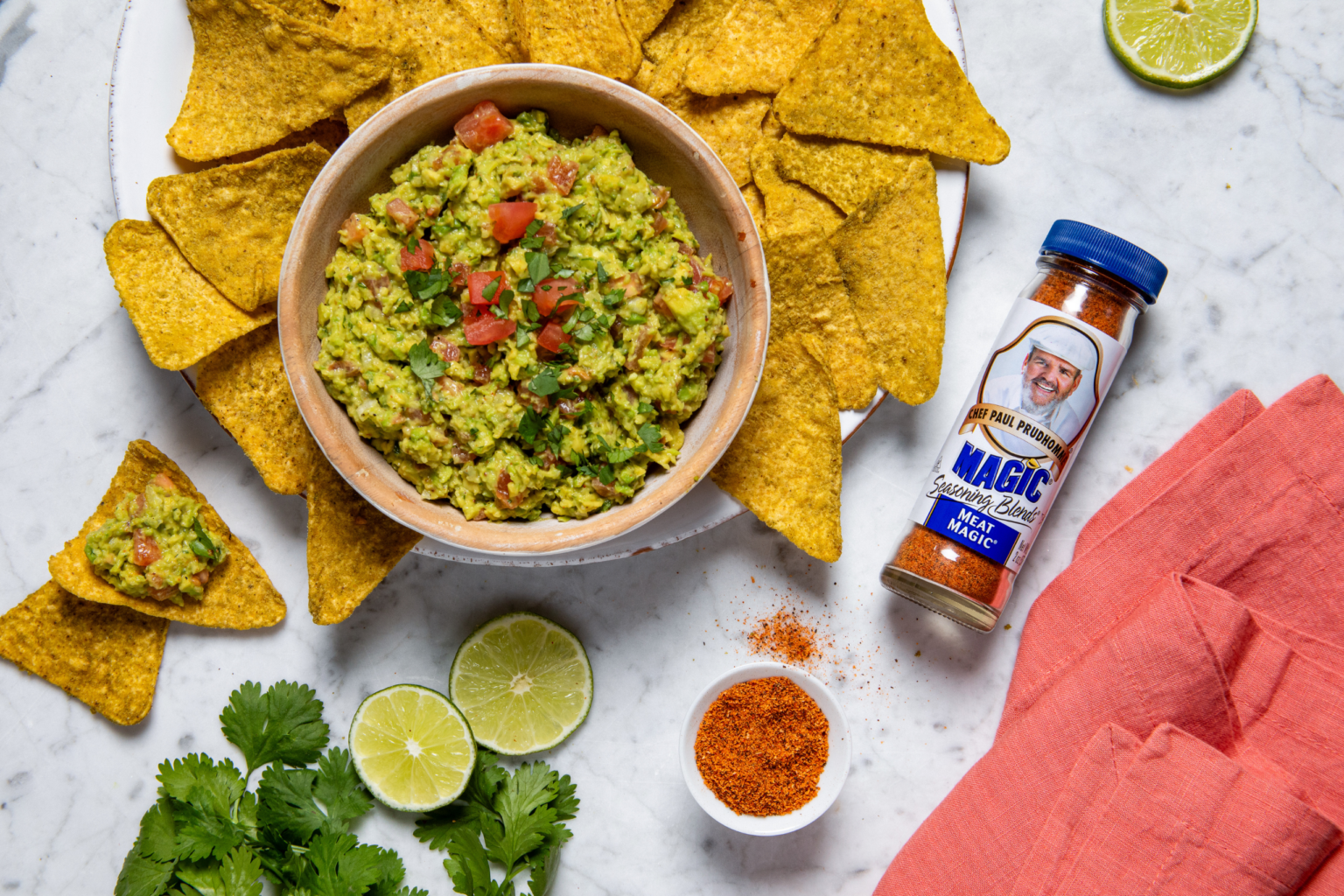 A bowl of guacamole garnished with diced tomatoes and cilantro is surrounded by tortilla chips. Nearby are lime halves, spice powder, and a red napkin.