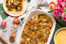 A casserole dish with baked seafood stuffing is surrounded by a seasoning bottle, bowls of sauce, and bright pink flowers on a white and green cloth.