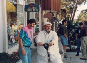 Chef Paul Prudhomme sits on a small motorized scooter outside an art gallery while artist George Rodrigue stands beside him, smiling, with Blue Dog paintings visible in the gallery window behind them.