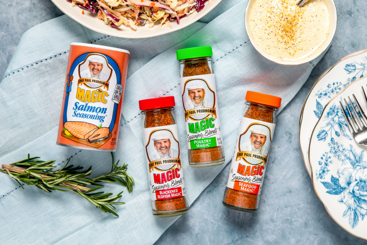 Four seasoning bottles and one can of salmon seasoning are arranged on a dark grey surface. Each container features a chef's image and colorful labels.