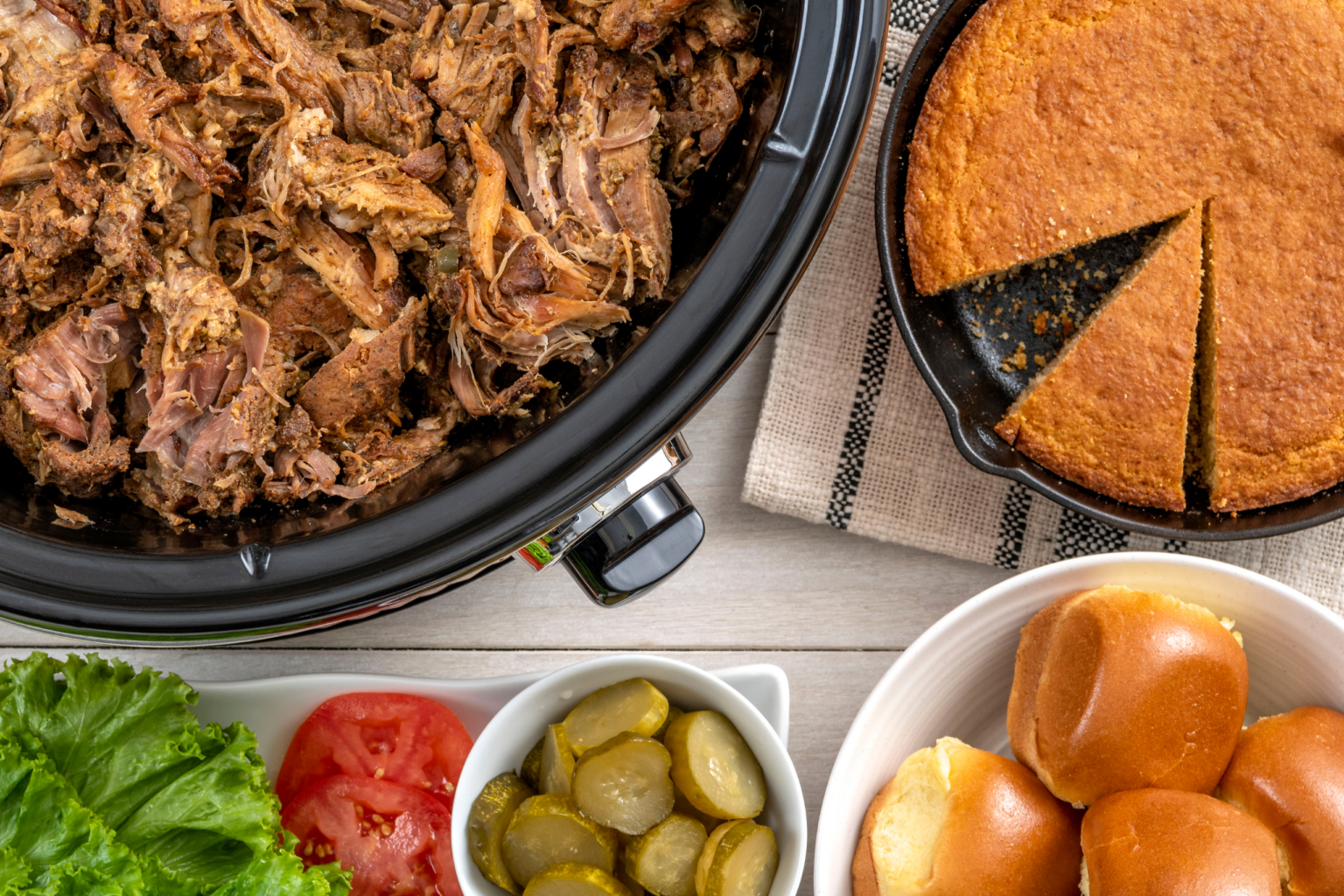 Slow-cooked pulled pork served with cornbread, slider buns, lettuce, tomatoes, and pickles for an easy summer cookout spread.