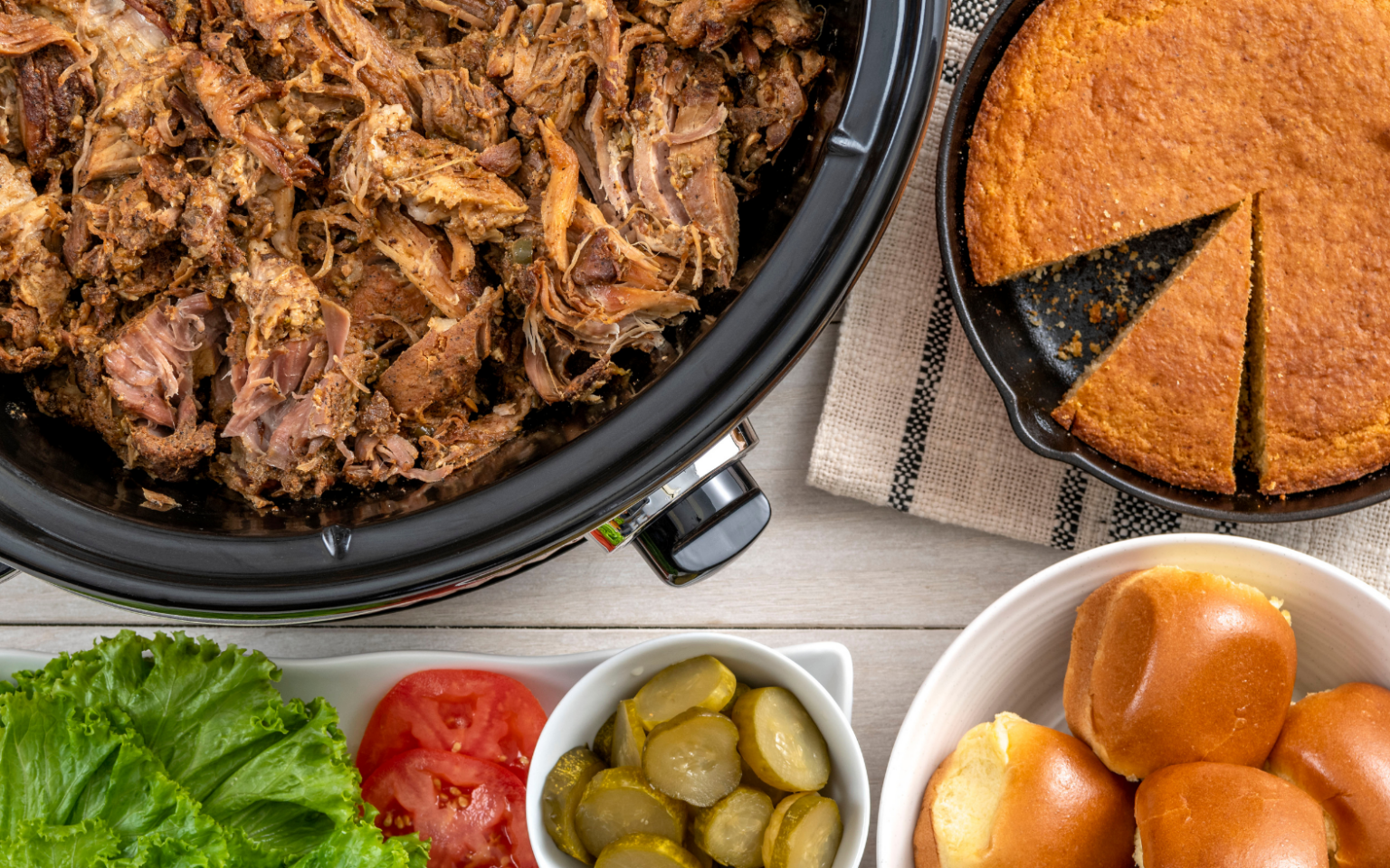 Slow-cooked pulled pork served with cornbread, slider buns, lettuce, tomatoes, and pickles for an easy summer cookout spread.