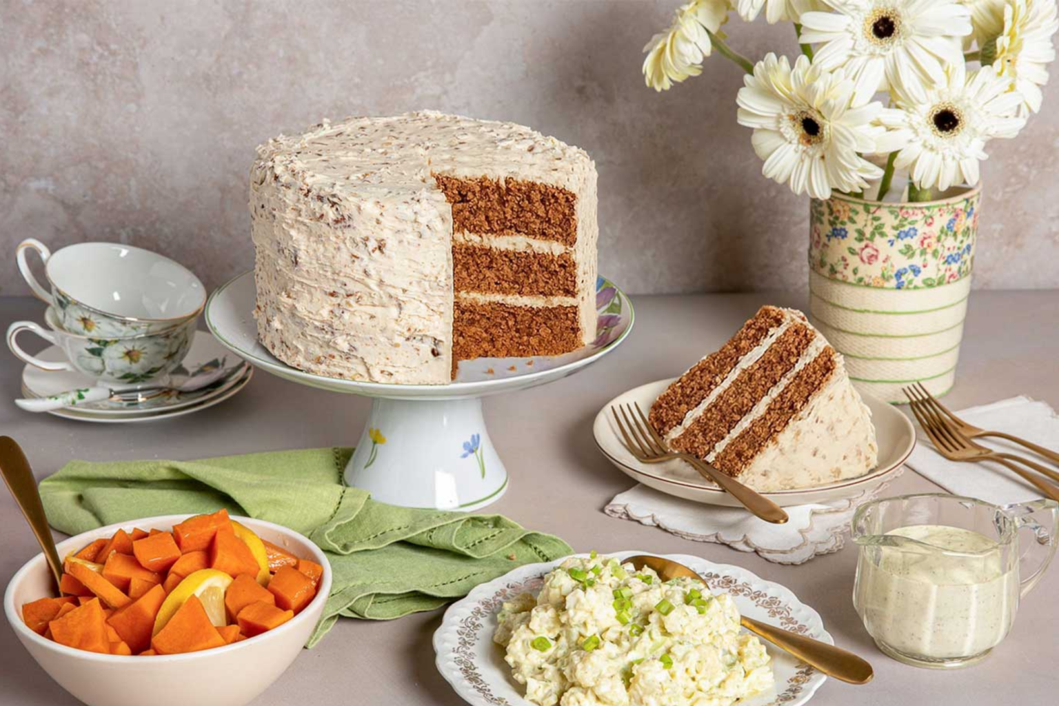 A three-layer cake with cream frosting on a stand, a slice on a plate, surrounded by bowls of fruit, salad, cups, and vibrant daisies in a vase.