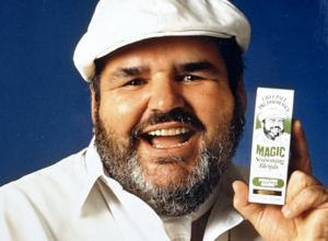Chef Paul smiling in a cap and white shirt holding a box of "Magic Seasoning Blends" against a blue background.
