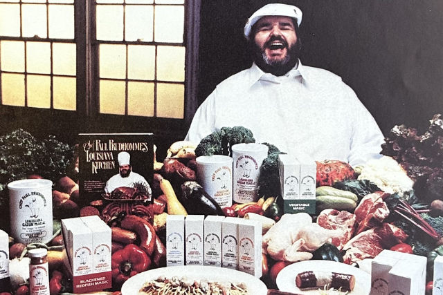 Chef Paul Prudhomme surrounded by fresh ingredients, seasoning products, and a cookbook.