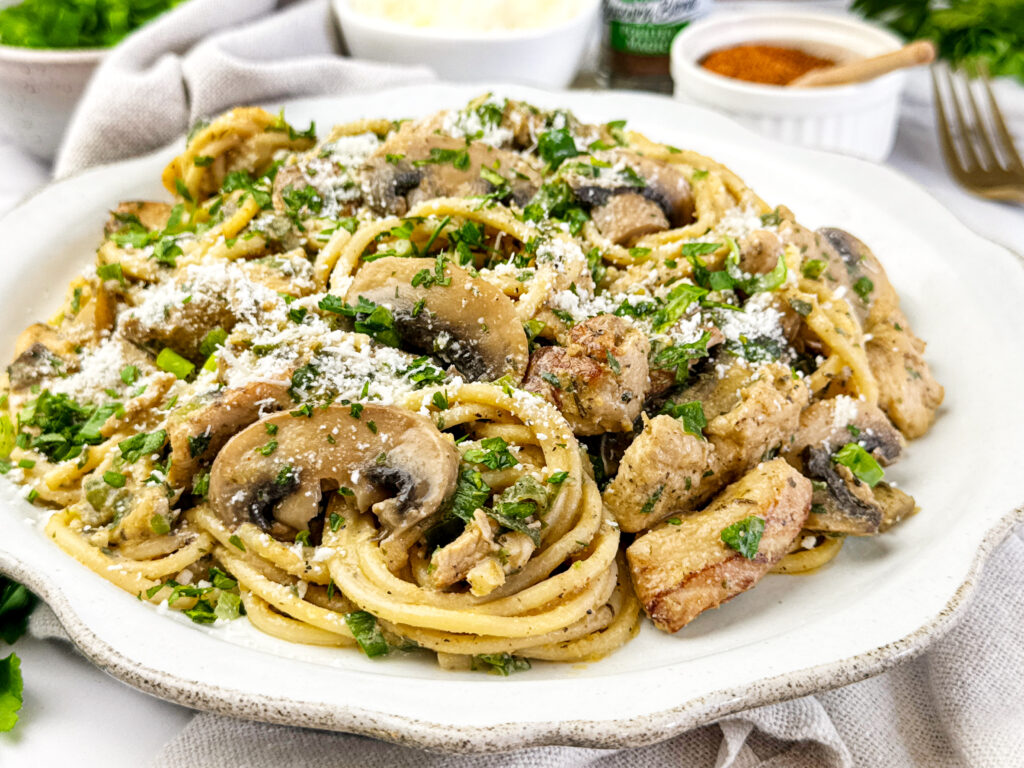 Chicken Diana pasta with fettuccine, sautéed mushrooms, and creamy sauce topped with fresh parsley and grated cheese on a white plate.