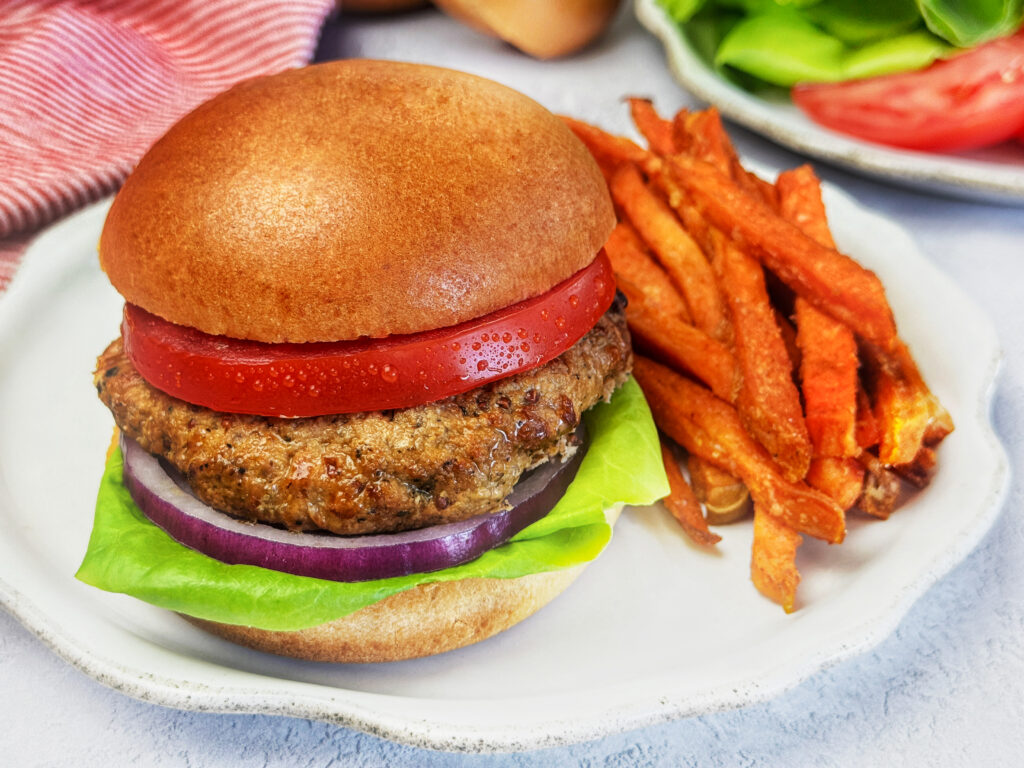 Turkey burger on a toasted bun topped with fresh lettuce, tomato slices, and red onion, served with sweet potato fries on a white plate.