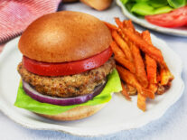 Turkey burger on a toasted bun topped with fresh lettuce, tomato slices, and red onion, served with sweet potato fries on a white plate.