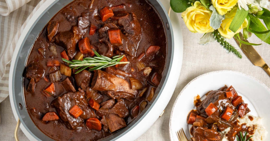 Slow cooker beef short ribs with carrots and onions in a rich sauce, garnished with fresh rosemary and served over mashed potatoes.