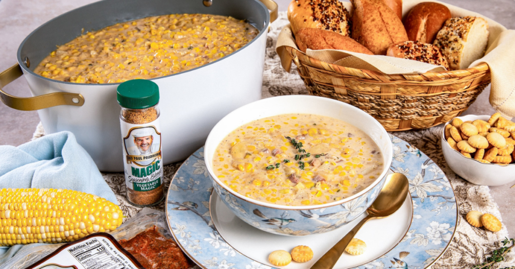 Slow cooker corn chowder served in a bowl with fresh thyme, corn on the cob, rolls, crackers, and Chef Paul Prudhomme’s Vegetable Magic seasoning.
