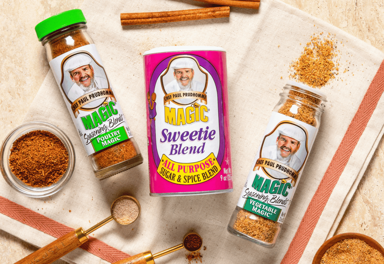 Chef Paul Prudhomme’s Poultry Magic, Sweetie Blend and Vegetable Magic arranged on a kitchen towel with spices and measuring spoons.